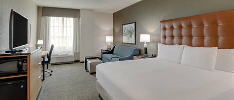 A hotel room with a large bed, a desk with a flat-screen TV, a chair, a small table, and two bedside lamps.