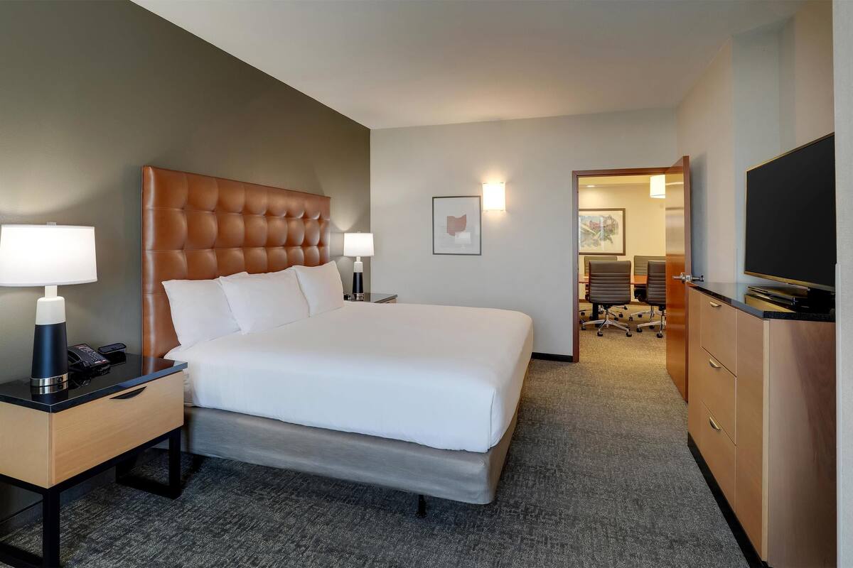 Ein Hotelzimmer mit einem großen Bett, Nachttischen mit Lampen, einem Fernseher und einem Schreibtisch mit Schubladen.