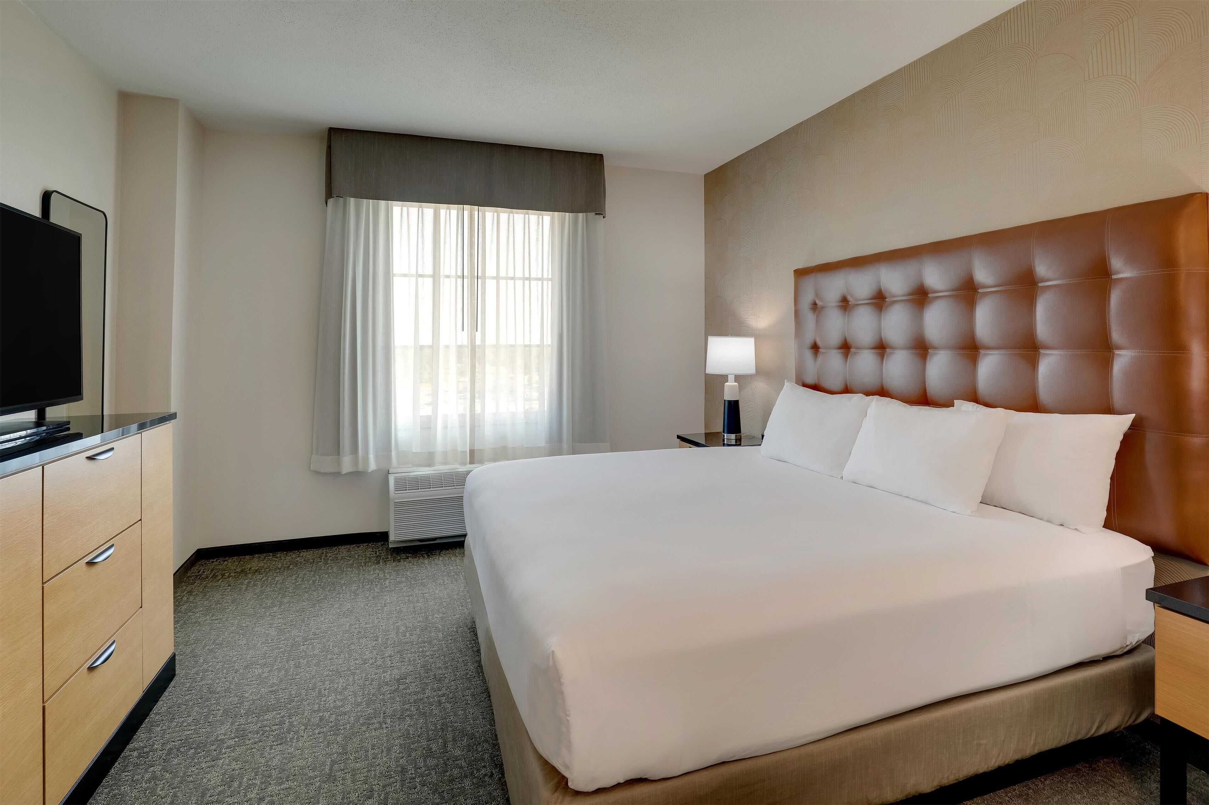 A hotel room with a large bed, a flat-screen TV, a bedside table with a lamp, and a window with sheer curtains.
