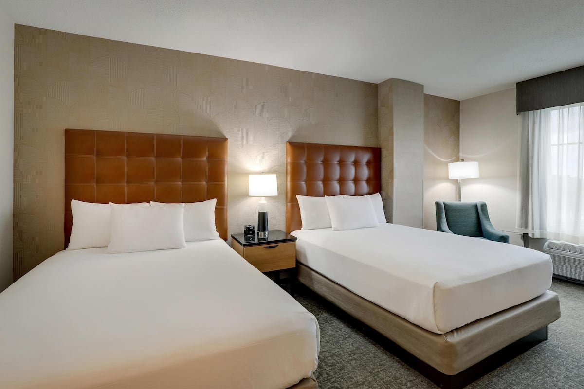 Una habitación de hotel con dos camas, una mesita de noche con lámpara, una silla y un gran ventanal con cortinas.