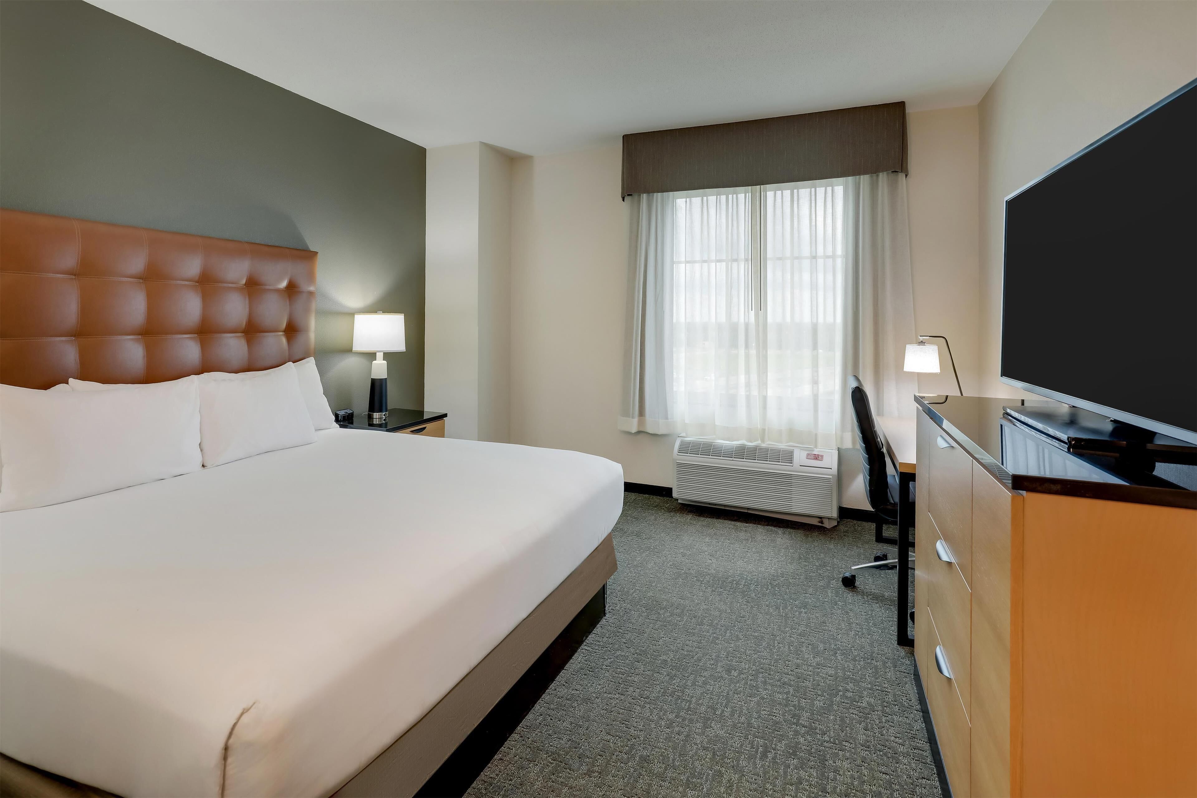 A hotel room with a large bed, a desk with a computer, a flat-screen TV, a window with curtains, and a wall-mounted air conditioner.