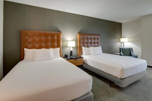 Habitación de hotel con dos camas, cada una con cabecera, mesita de noche con lámpara y una silla.
