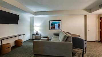 A hotel room with a sofa, a small table, a lamp, a TV mounted on the wall, and a framed picture.
