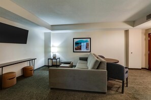 A hotel room with a sofa, a small table, a lamp, a TV mounted on the wall, and a framed picture.