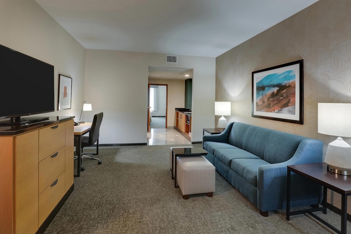 A hotel room with a TV, a desk, a blue sofa, a small ottoman, and a framed picture on the wall.