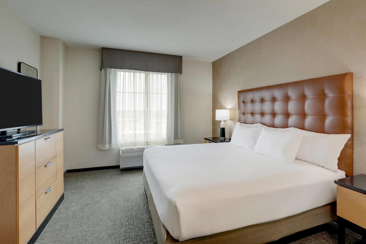 Habitación de hotel con una cama grande, un televisor sobre una cómoda de madera, una mesita de noche con lámpara y una ventana con cortinas.