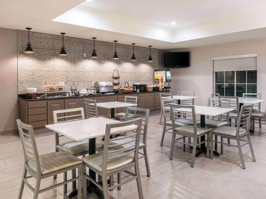Uma zona de refeições moderna com várias mesas e cadeiras, um balcão de cozinha com eletrodomésticos e uma televisão de ecrã plano na parede.