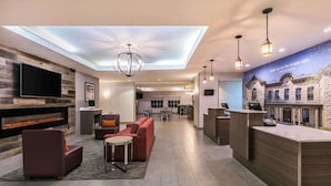 A modern hotel lobby with a flat-screen TV, a fireplace, a reception desk, and a mural of a building. - La Quinta Inn & Suites by Wyndham Fredericksburg (Fredericksburg)