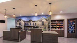 A reception area with two wooden reception desks, overhead pendant lighting, a mural of a historic building, and shelves stocked with snacks and beverages. - La Quinta Inn & Suites by Wyndham Fredericksburg (Fredericksburg)