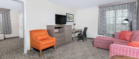 A hotel room with a grey carpet, orange armchair, grey desk, black office chair, pink sofa, and a TV on the desk.