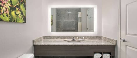 Une salle de bain moderne avec un lavabo doté d’un plan de travail en granit, un grand miroir et un porte-serviettes où sont soigneusement pliées les serviettes.