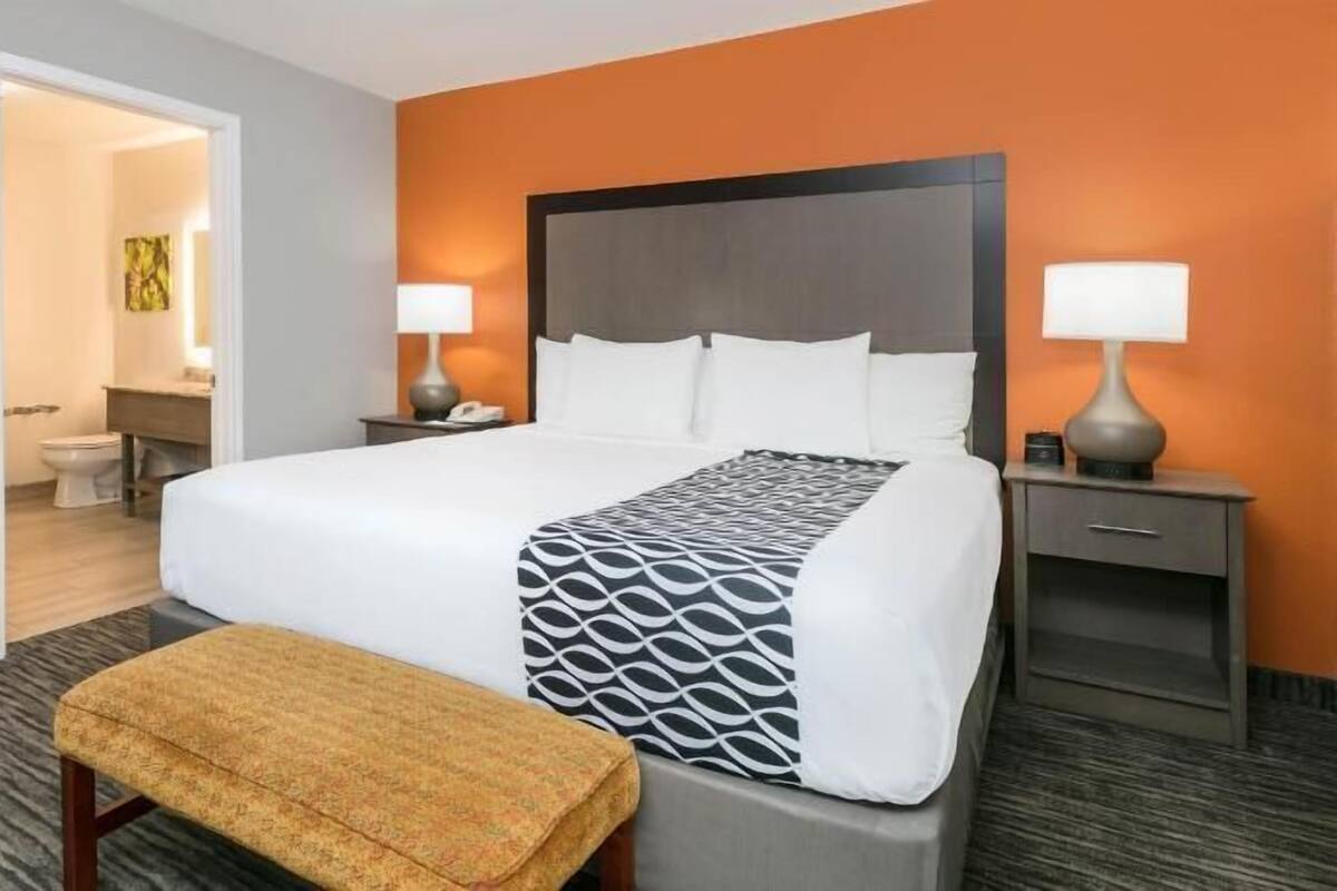 Habitación de hotel con una cama grande, una mesita de noche con lámpara, un pequeño banco y un baño visible a través de una puerta abierta.