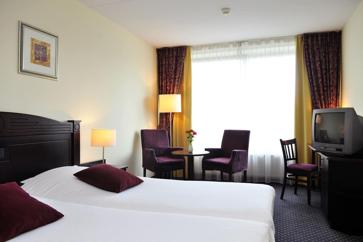 Une chambre d’hôtel comprenant un lit, deux fauteuils, une petite table avec une lampe, un téléviseur et une fenêtre avec des rideaux.