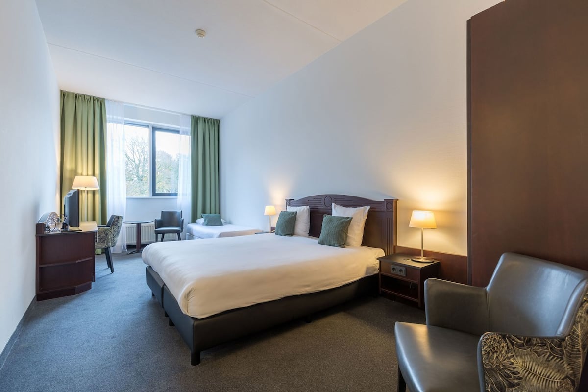 Une chambre d’hôtel avec un grand lit, deux tables de chevet, un bureau avec une chaise, une fenêtre avec des rideaux et une chaise agrémentée d’un coussin à motifs.
