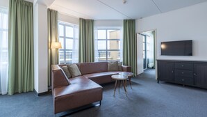 A modern hotel room with a leather sofa, two small round tables, a flat-screen TV, and a large window with curtains.