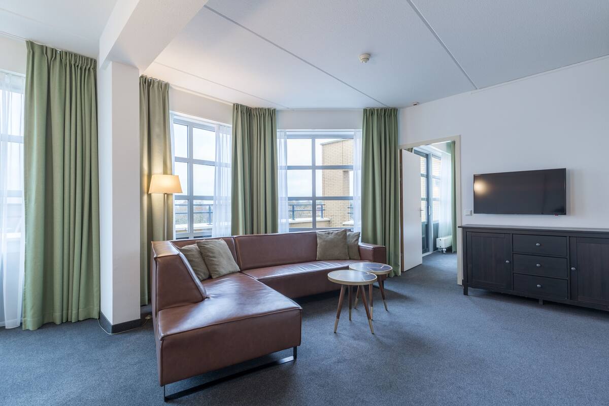 Une chambre d’hôtel moderne avec un canapé en cuir, deux petites tables rondes, un téléviseur à écran plat et une grande fenêtre avec des rideaux.