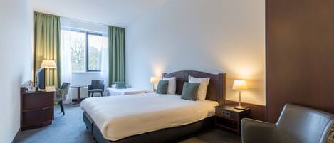 A hotel room with a large bed, two bedside tables with lamps, a desk with a chair, a small table with a chair, and a window with curtains.