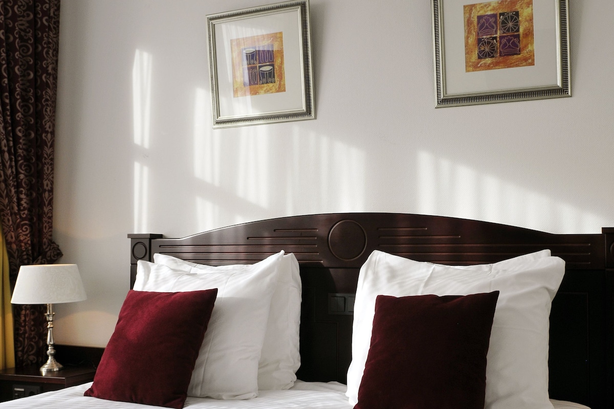 A hotel room with a large bed, two framed pictures on the wall, a lamp, and a patterned throw blanket.