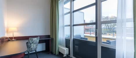 A hotel room with a large window, a desk with a lamp, a chair, and a radiator.