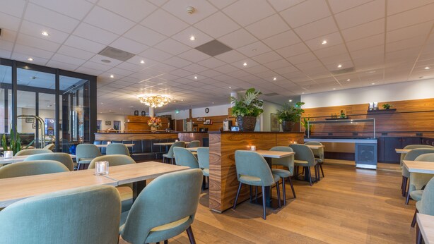 A modern restaurant with wooden flooring, grey upholstered chairs, and a central island with plants.