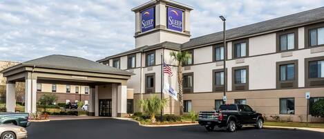 A hotel with a central tower displaying the name "Sleep Inn & Suites," a parking lot with several vehicles, and a well-maintained entrance area.