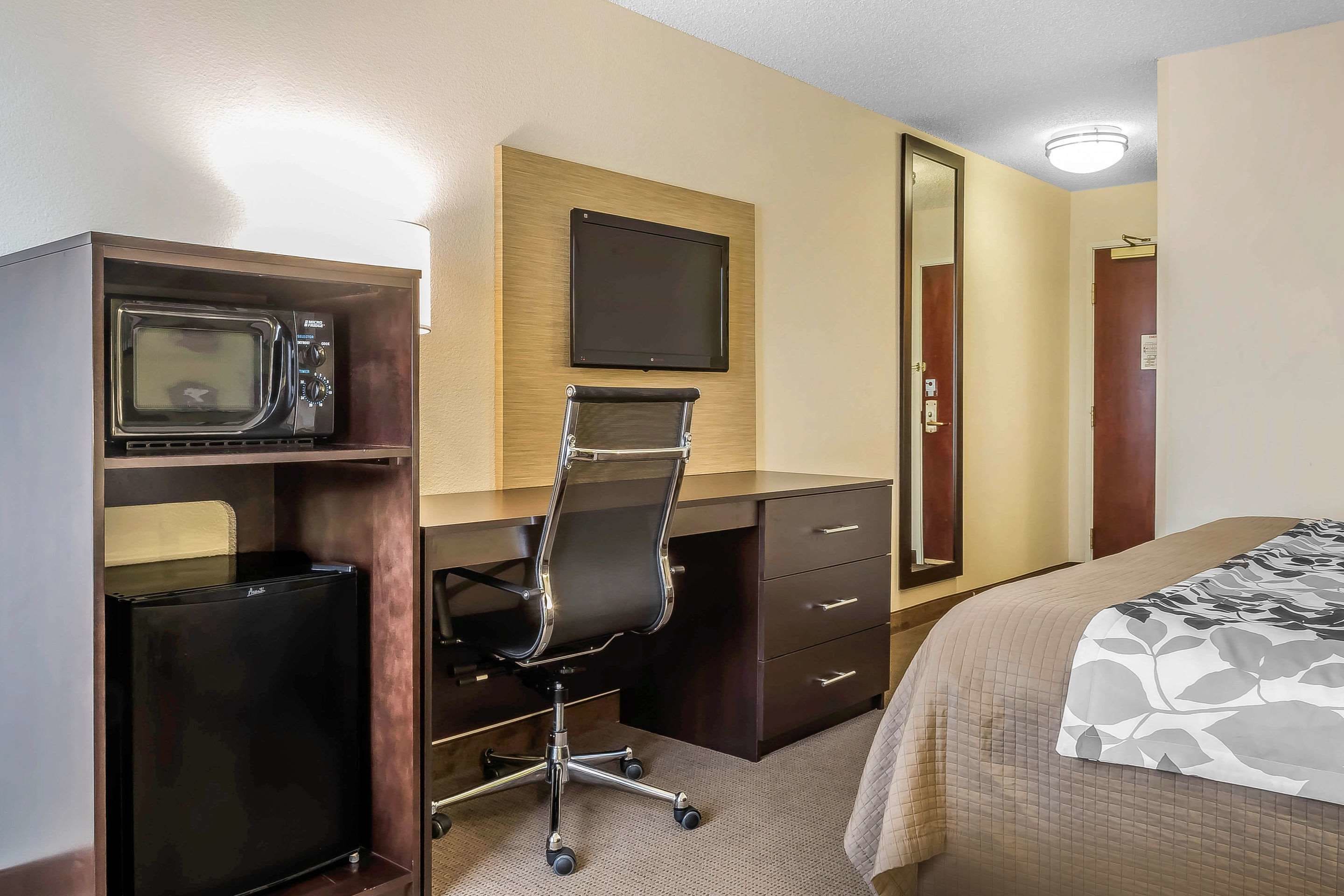 Quarto de hotel com cama, escrivaninha com computador, mini-geladeira, micro-ondas, cadeira, televisão e porta.