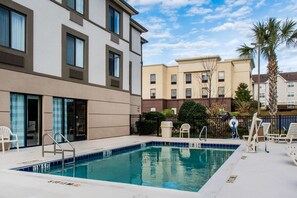 Una piscina con una escalera metálica, rodeada de sillas de descanso y una palmera.
