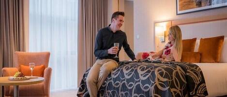 Una habitación de hotel con una cama, una silla con una bandeja de fruta y una copa de vino, y un cuadro en la pared.