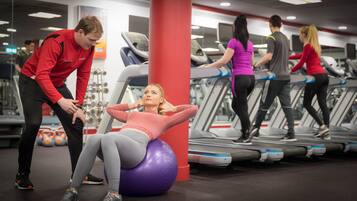 Een sportschool met loopbanden, een persoon die een fitnessbal gebruikt en een andere persoon die assisteert.