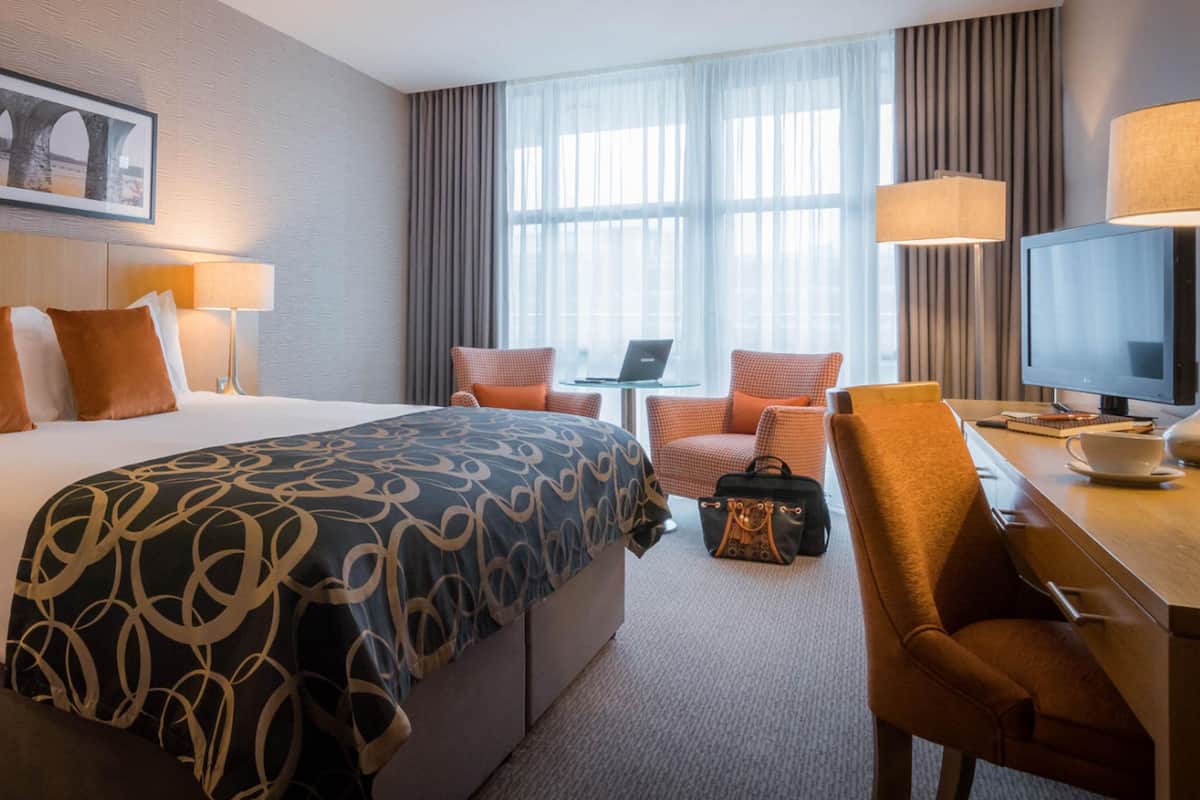 Ein Hotelzimmer mit einem großen Bett, zwei Sesseln, einem Schreibtisch, einem Fernseher und einem Laptop.