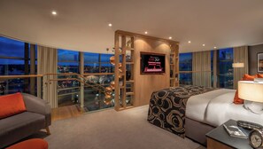 Habitación de hotel con una cama grande, un escritorio, una silla y vistas a la ciudad al atardecer.