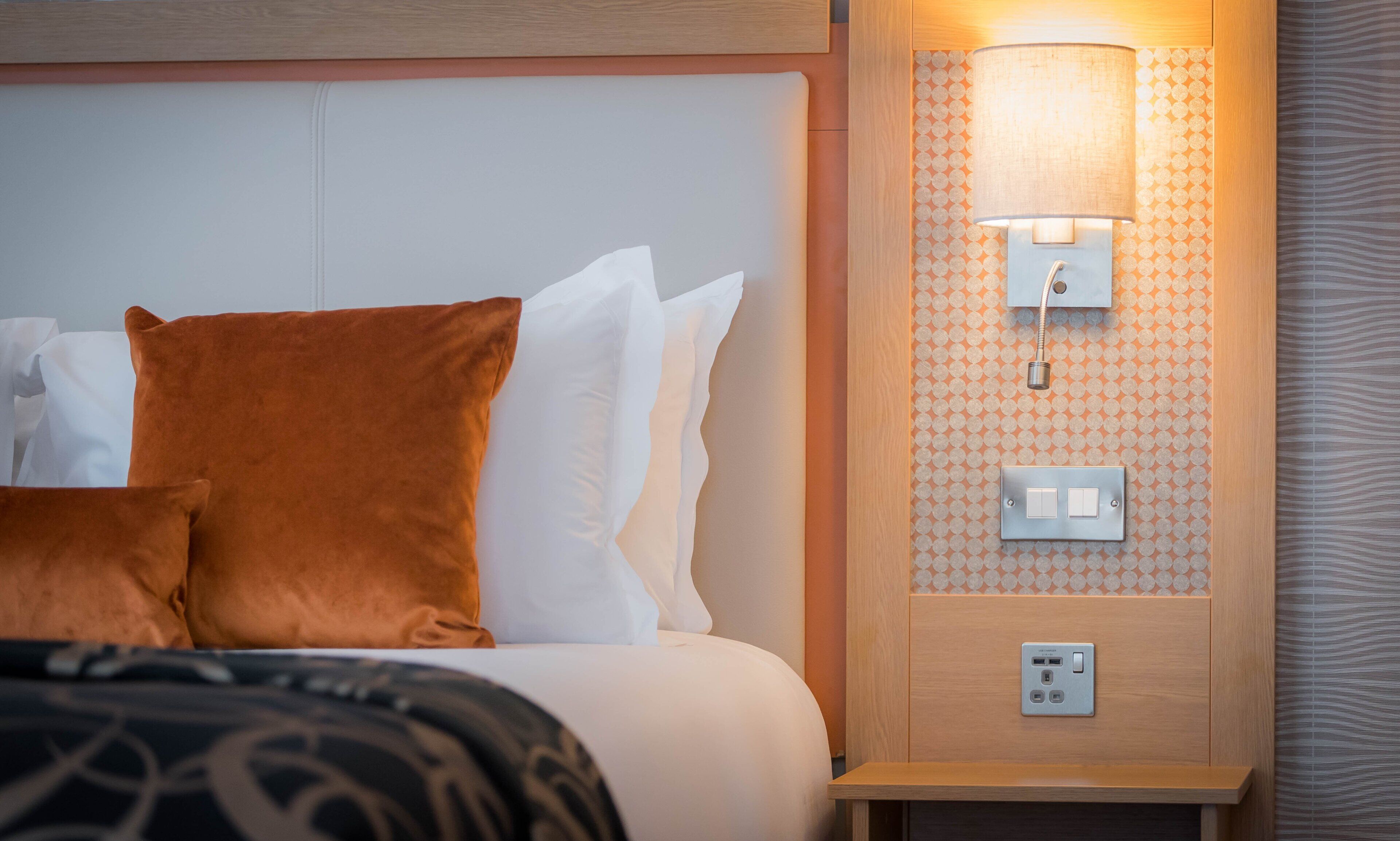 Een net opgemaakt bed met witte en bruine kussens, een bedlampje en een stopcontact aan de muur.