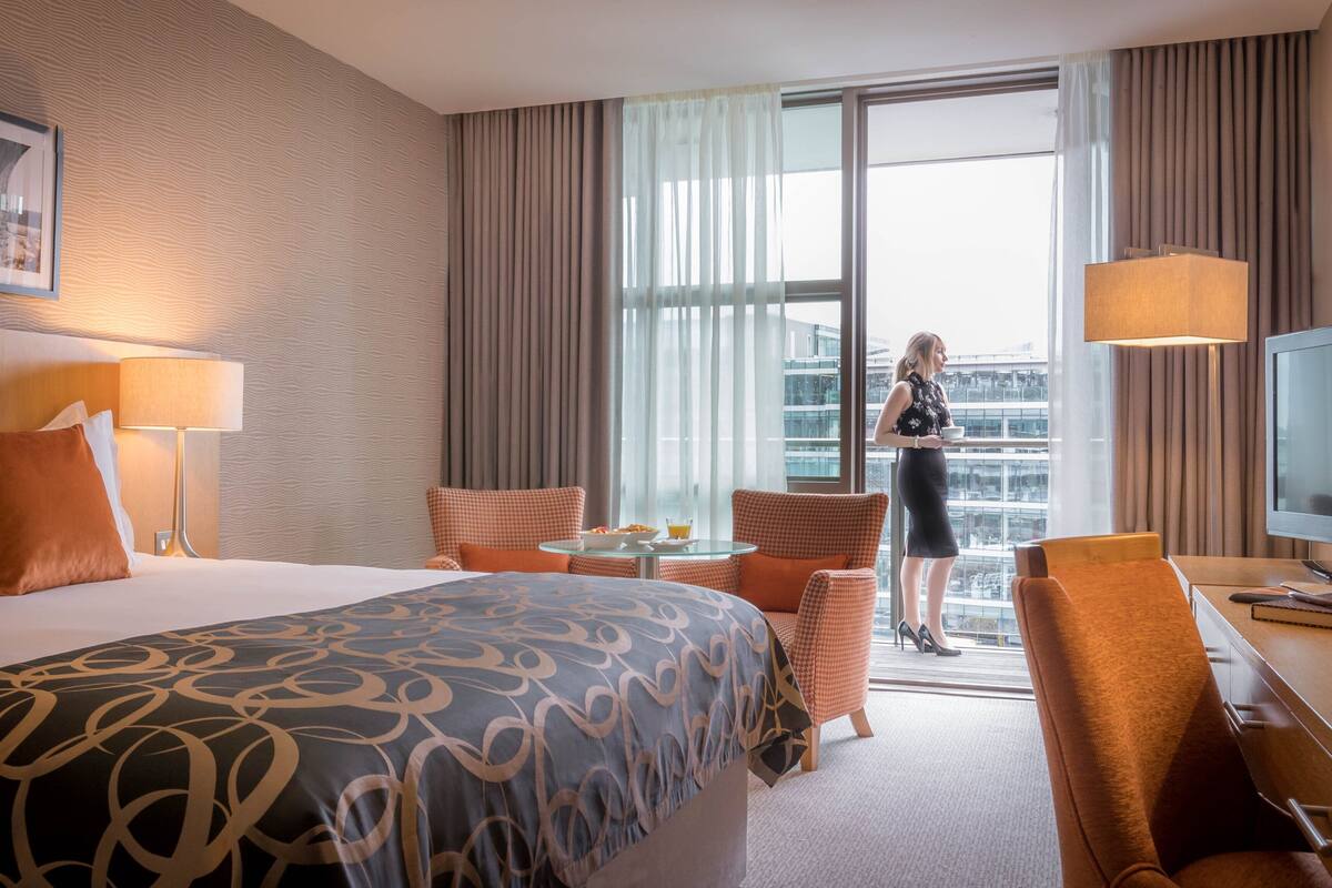 Ein Hotelzimmer mit einem großen Bett, zwei Sesseln, einem kleinen Tisch, einem Schreibtisch, einem Fernseher und einem Fenster mit Blick auf die Stadt.