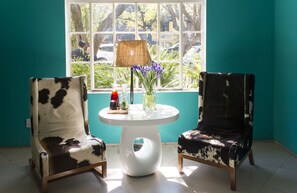 A room with teal walls, a window with a view of parked cars and trees, two cowhide patterned chairs, a white table with a lamp, and a vase with purple flowers.