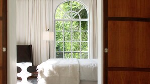 Quarto com uma grande janela em arco, uma cama com cobertor branco, mesa de cabeceira e abajur.