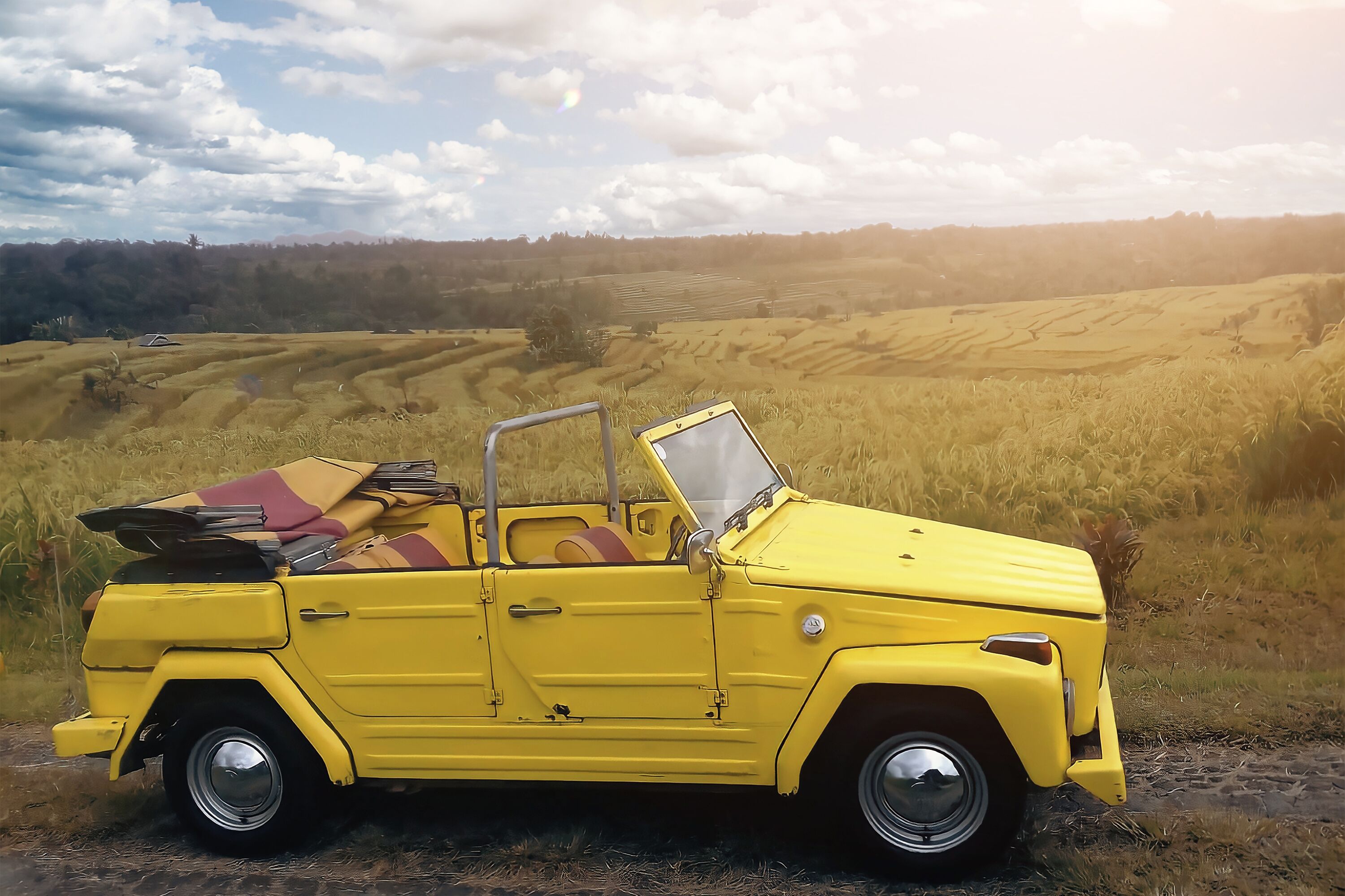 A yellow convertible car parked on a dirt path with a scenic view of terraced fields and mountains in the background.