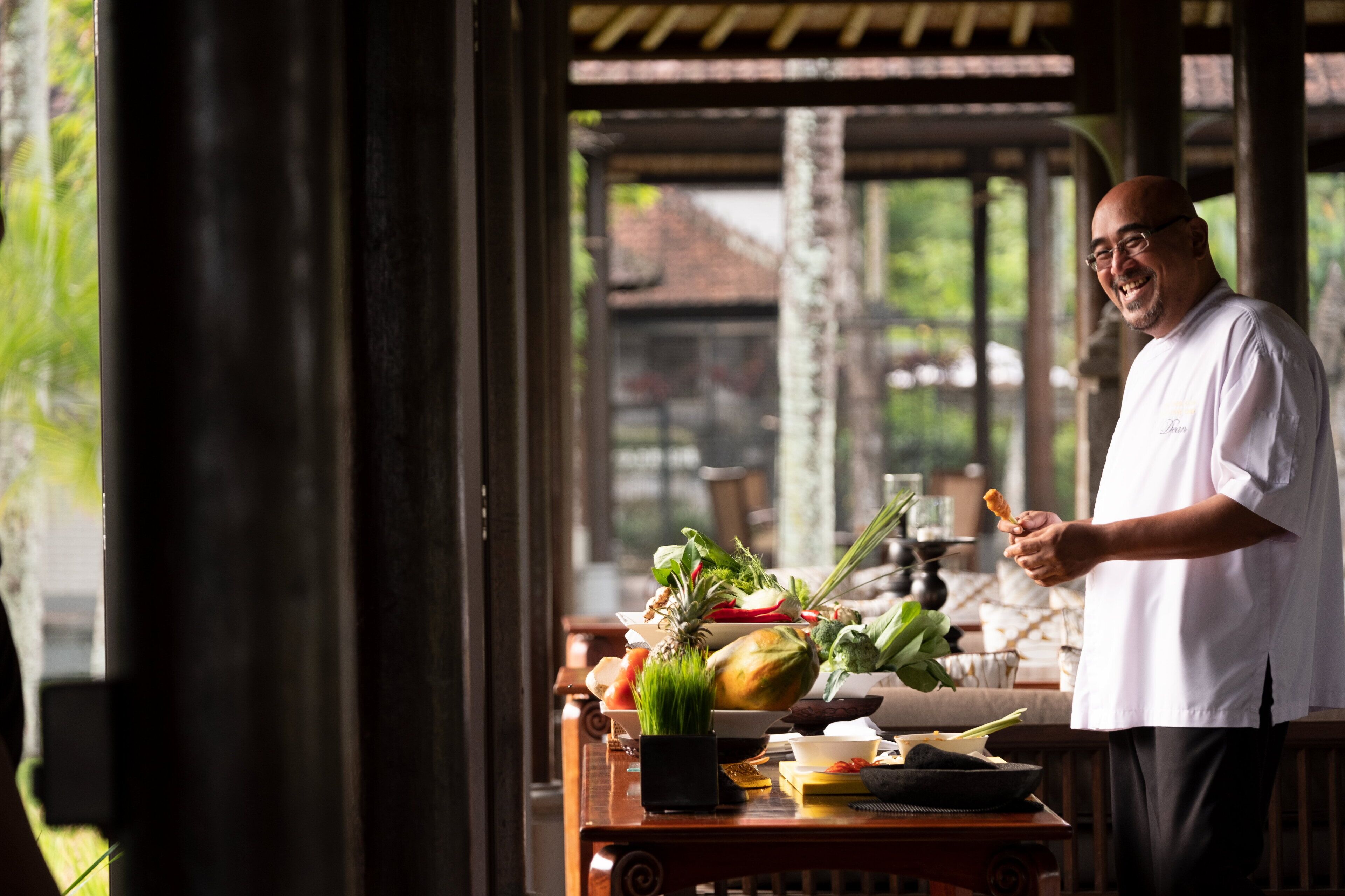 A chef standing in a covered outdoor area with a wooden table displaying fresh vegetables and cooking utensils.
