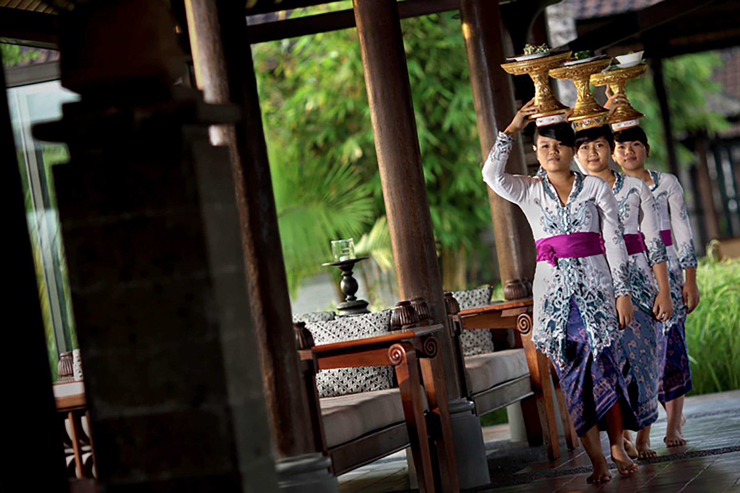 A traditional Balinese ceremony with participants carrying ornate golden vessels, set in a wooden pavilion with a tiled floor and a view of lush greenery.