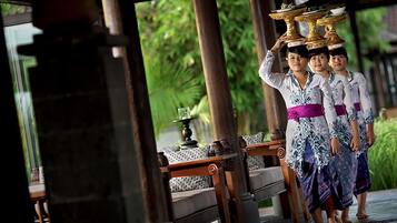 A traditional Balinese ceremony with participants carrying ornate golden vessels, set in a wooden pavilion with a tiled floor and a view of lush greenery.