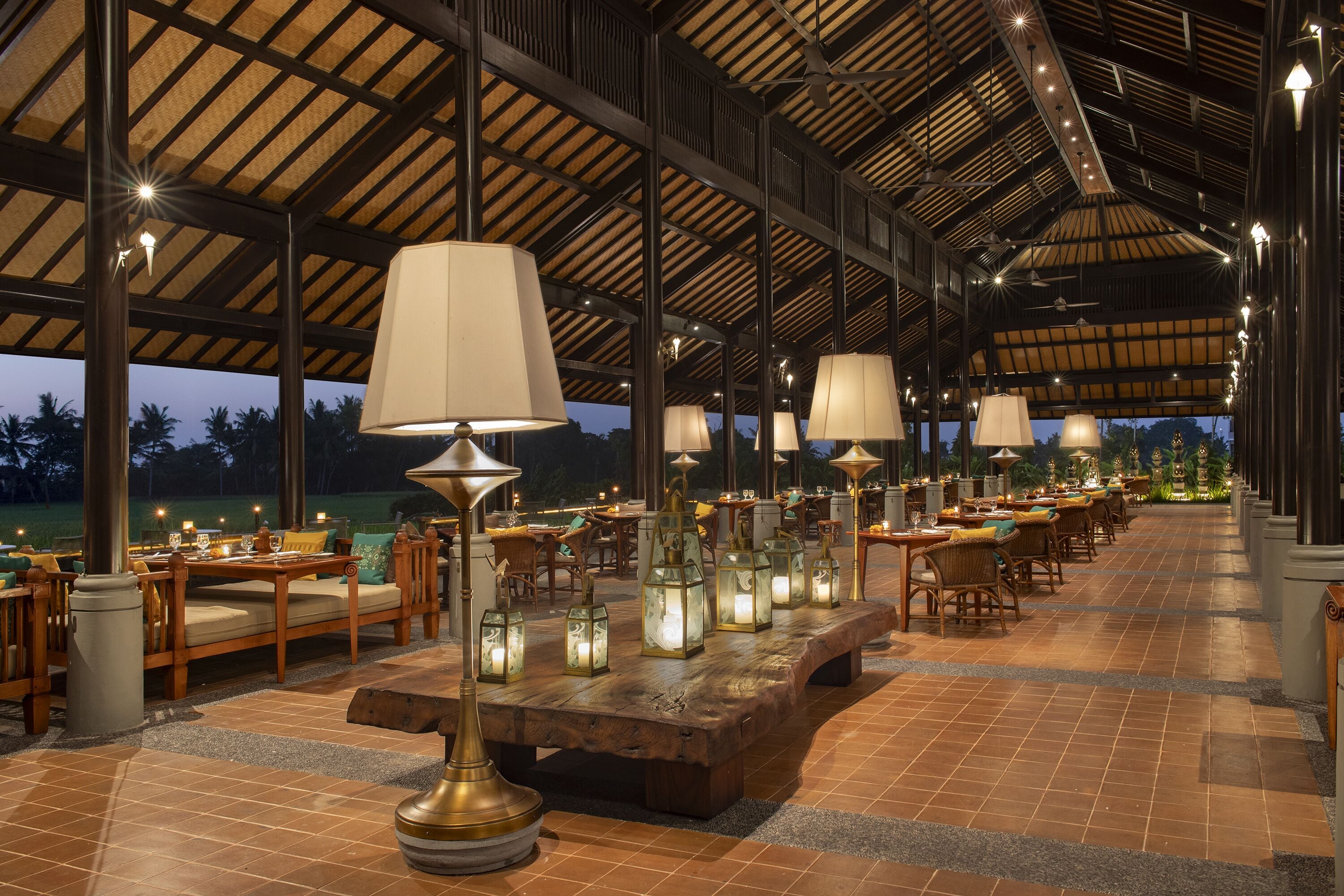 An outdoor dining area with wooden tables and chairs, illuminated by hanging lamps and lanterns.