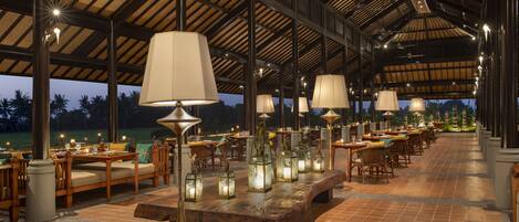 An outdoor dining area with wooden tables and chairs, illuminated by hanging lamps and lanterns.