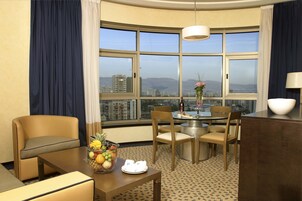 A hotel room with a dining area, a large window with a city view, and a fruit bowl on the table.