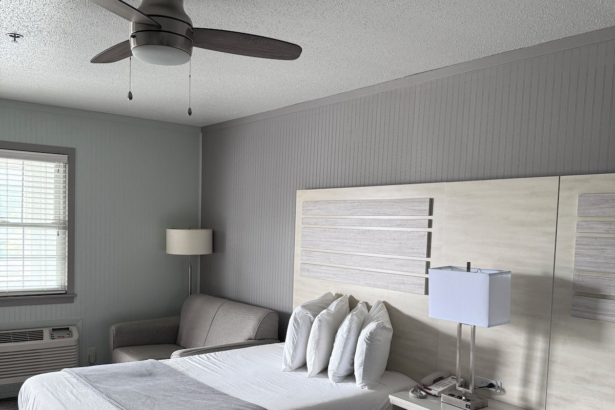 A hotel room with a large bed, a smaller bed, a desk with a lamp, a chair, a ceiling fan, and a window with blinds.