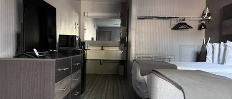A hotel room with a bed, a desk with a computer, a wardrobe, a ceiling fan, and a bathroom visible through an open door.