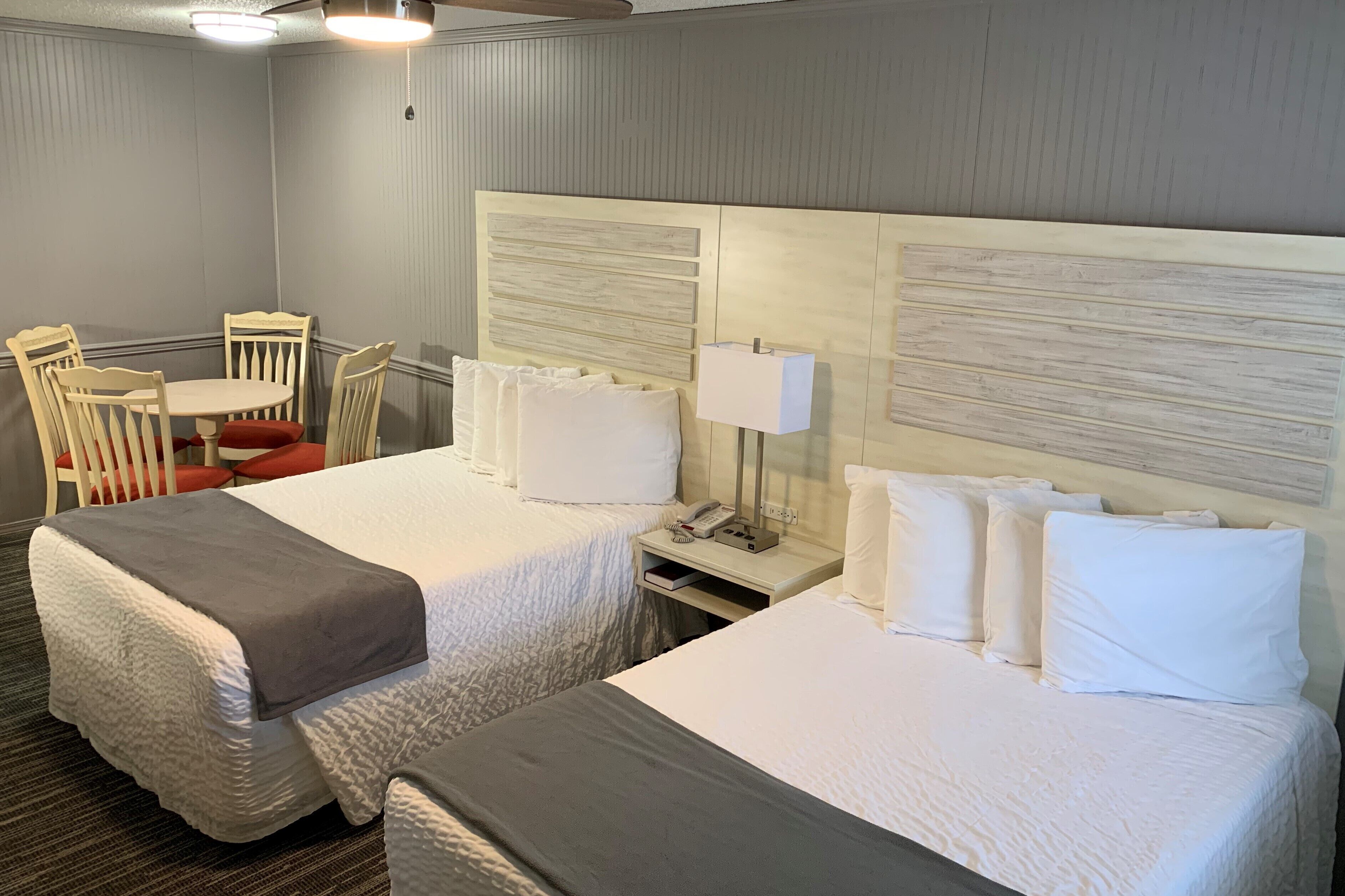 A hotel room with two beds, a small table, chairs, a lamp, and a telephone on the nightstand.