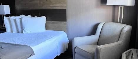 A hotel room with a bed, a chair, a lamp, and a heater.