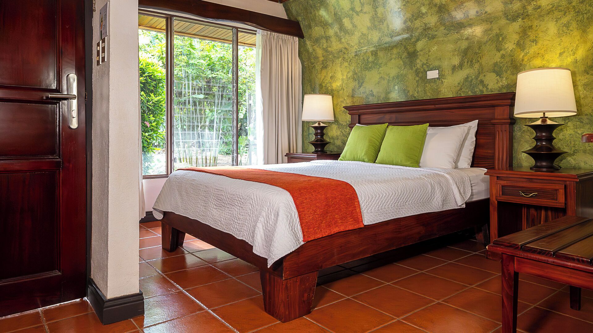 Une chambre avec un grand lit, deux tables de chevet avec des lampes, une porte en bois et une fenêtre avec des rideaux.
