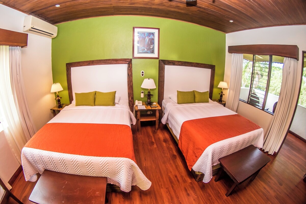 A hotel room with two beds, a wooden floor, a green wall, and a window with curtains.