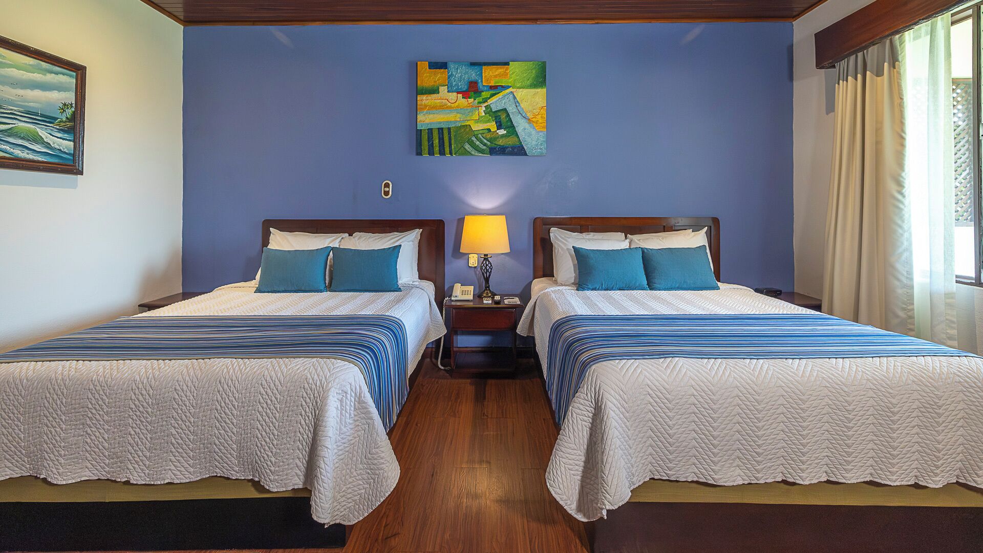 A hotel room with two beds, each with a quilted bedspread and pillows, a nightstand with a lamp, and a painting on the wall.