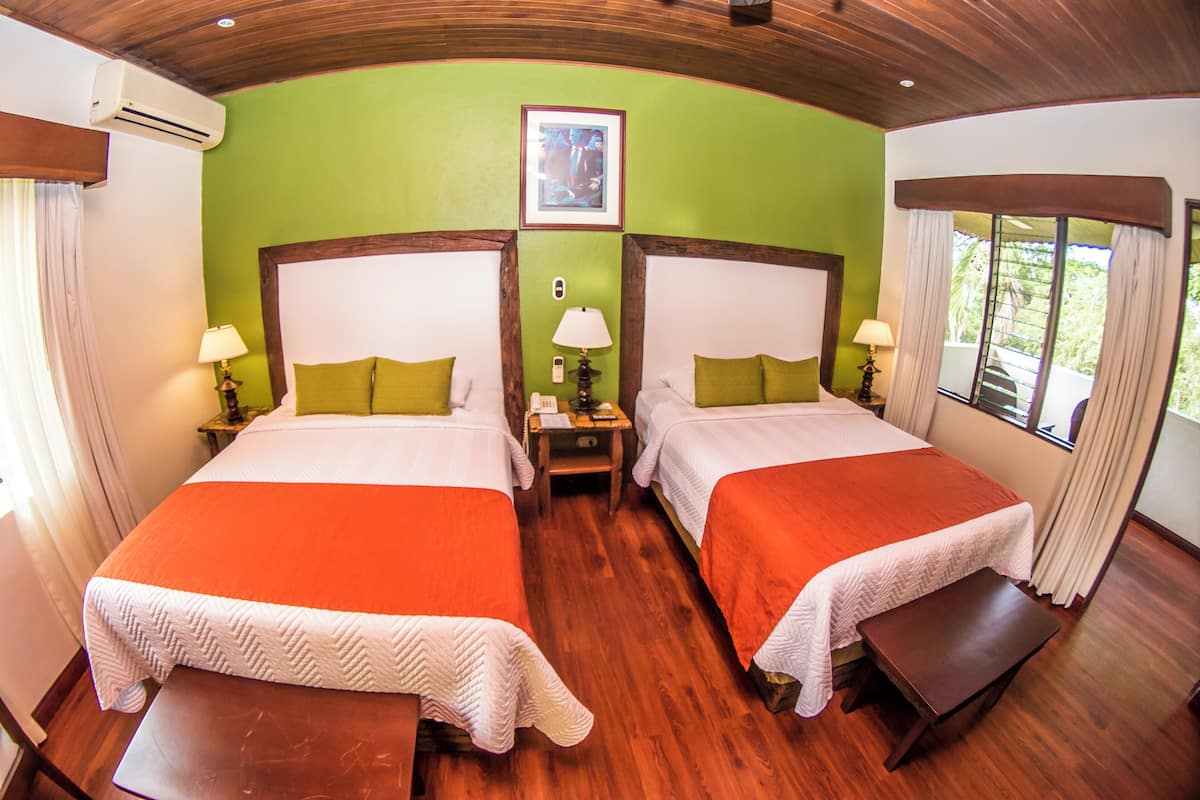 A hotel room with two beds, a wooden floor, a green wall, and a window with curtains.
